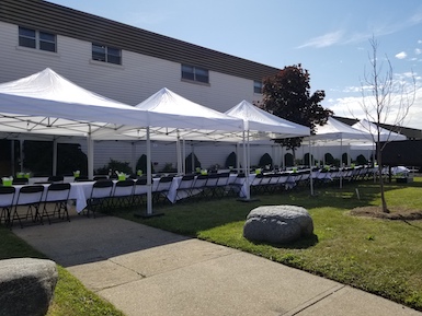 5 10 foot by 10 foot tents set up in a row with 8 foot by 30 inch tables set up under the tents with 60 inch by 126 inch white tablecloths and black plastic and metal folding chairs on 2 sides of the tables 
