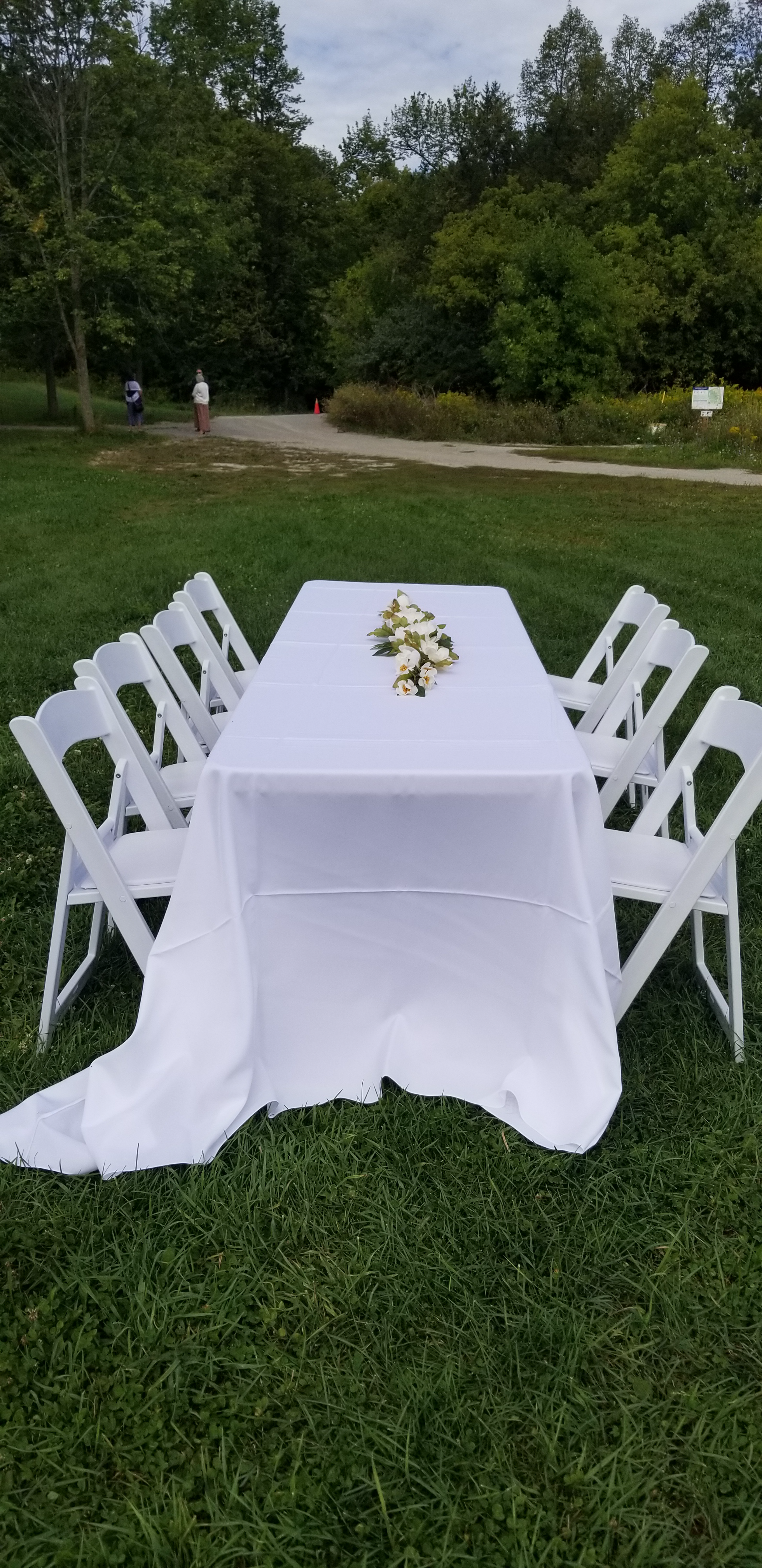 8 foot by 30 inch table with 90 inch by 156 inch white tablecloth on it, a flower arrangement and 4 of the white resin chairs with padded seats on 2 sides of the table