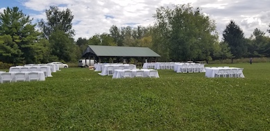 wedding party set up in Picnic Area of the Kortright Centre in Vaughan there are 12 8 foot by 30 inch tables set up in 3 rows of 4 tables each with white tablecloths and white resin chairs with padded seats set up on 2 sides of tables