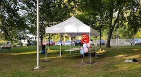 10 foot by 20 foot white pop up tent set up with weights on the grass in the Beaches neighbourhood of Toronto with tables underneath the tent and a bunch of baloons including the number 6 balloon in red and 5 other balloons 3 in pink and 2 in baby blue near the tent 