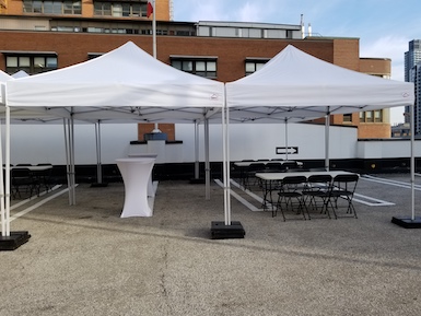 2 10 by 10 white pop up tents set up with tent weights on top level of downtown Toronto parking lot with 2 cruiser tables with white stretch tablecloths under one tent and 2 6ft by 30 inch folding tables with 6 black plastic with metal folding chairs at each table under the second tent